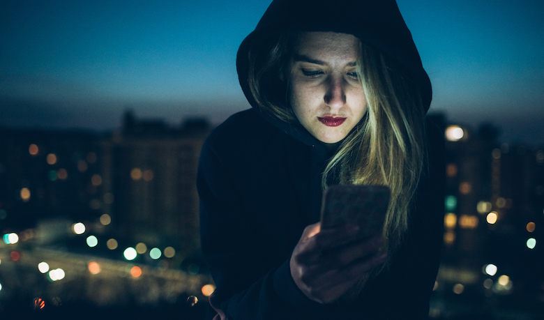 Young woman sitting on rooftop at night, using smartphone, illuminating face
